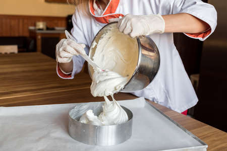 Female chef cook in rubber gloves pours the vanilla cake-mix dough into form for making cake. Young...
