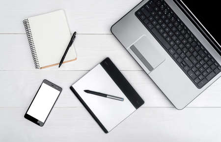 Top view on white wooden table with open blank laptop computer, cell phone, empty diary and graphic...