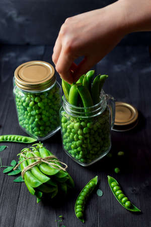 Two full glass jars with fresh mature green peas near bunch of pods tied with rope on black wooden background. Bio healthy food. Woman's hand taking green pea pod from jarの写真素材