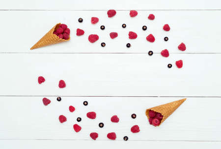 Frame of sweet fresh raspberries and blackcurrants in waffle cones, free space. Fresh berries in cones on white wooden background with blank space. Summer and healthy food concept flat lay, top viewの写真素材