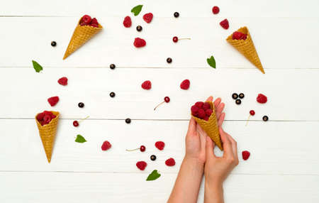 Top view of sweet fresh organic raspberries in waffle cones and one cone in girl's hands. Fresh berries in cones and on white wooden background. Summer and healthy food concept の写真素材
