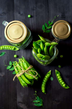 Two full glass jars with fresh mature green peas near bunch of pods tied with rope on black wooden background. Bio healthy food. Green peas, pods, pea leaves and jar on wooden tableの写真素材