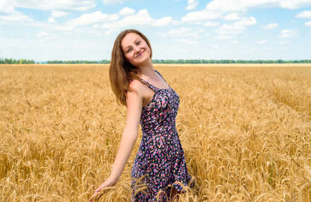 Beautiful young woman walking in golden wheat field with cloudy blue sky background, free space. Liberty, peace of mind concept. Girl in spikes of ripe wheat field under blue sky の写真素材