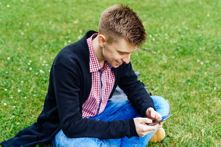 Portrait of young happy man sitting on green grass and using mobile smart phone outdoor in summer, copy spaceの写真素材