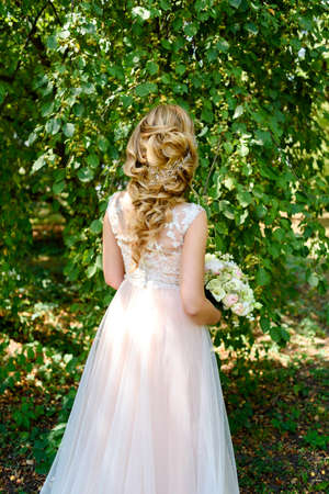 Beautiful bride in wedding dress with bridal bouquet in the park outdoors, back view. Blond girl with curly hair styling and jewelry. Wedding hairstyle for long hair with stylish hair accessoryの写真素材