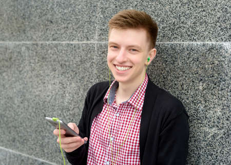 Cheerful happy young man in plaid shirt and jacket listening to music with headphones from cell...