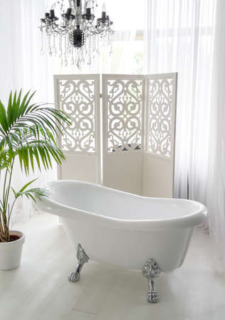 Beautiful luxury vintage empty bathtub near big window in bathroom interior, free space. Freestanding white bath near folding screen and palm tree, copy space の写真素材