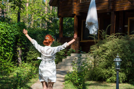 Beautiful redhead bride in silk dressing gown running with hands up to her wedding dress hanging on wooden house in the forest, free space. Wedding morning preparation in sunny summer dayの写真素材