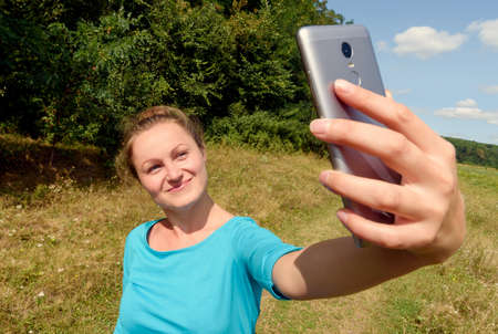 Portrait of smiling cheerful young woman making selfie photo on smartphone in park outdoors  at...