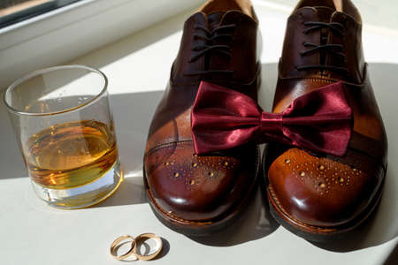 Close up of burgundy bowtie on brown leather man shoes, wedding rings and glass of cognac. Modern...