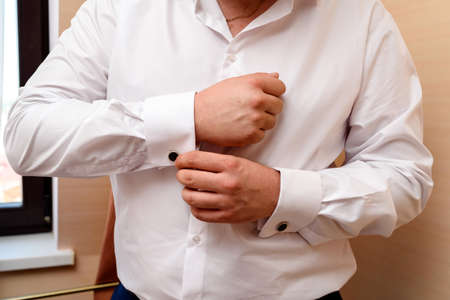 Man buttons cufflink on cuffs sleeves luxury white shirt. Cose up of man hand wears white shirt and...