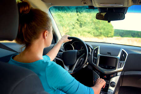 Young casual woman in blue dress driving a car, back view. Beautiful young girl at the wheel of car with black interior looking at the roadの写真素材