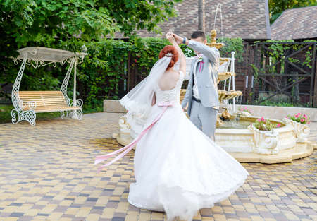 Happy groom and beautiful redhead bride in white dress holding hands and dancing outdoors, free space. Beautiful wedding couple spinning in wedding day. Wedding dress with bow on back flutters in the windの写真素材