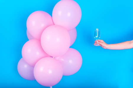 Girl hand holding wineglass with champagne near pink air balloons on blue background, free space. Concept of love, happiness, birthday, valentine, New Year party. Holiday pink balloonsの写真素材