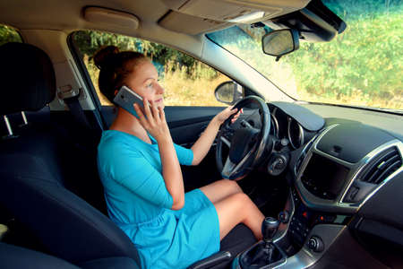 Side view of beautiful young woman driving a car and talking on smart phone, free space. Business woman sitting in car with cell phone,  looking on roadの写真素材
