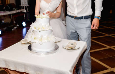 Stylish bride and groom cutting white multi level wedding cake with fresh flowers in the restaurant at wedding receptionの写真素材
