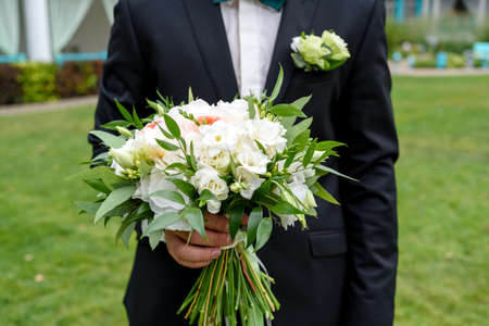 Bridal wedding bouquet of white flowers and greenery with ribbons in groom hand. Groom waiting for bride outdoor. Young handsome groom in black suit standing and holding wedding bouquetの写真素材