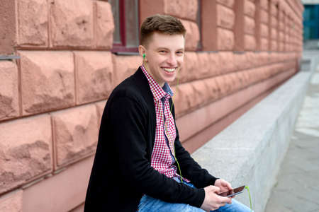 Cheerful happy young man in plaid shirt and jacket listening to music with headphones from cell phone and singing outdoors. Student with smart phone and headphonesの写真素材