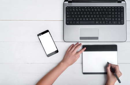 Top view of open laptop computer, mobile smart phone and girl's hands with graphic tablet, free space. Girl's hands with graphic tablet retouching image on laptop. Mobile phone with blank white screenの写真素材