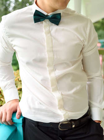 Close-up of businessman or groom in white shirt and bow-tie standing outdoors. Man wears white shirt...