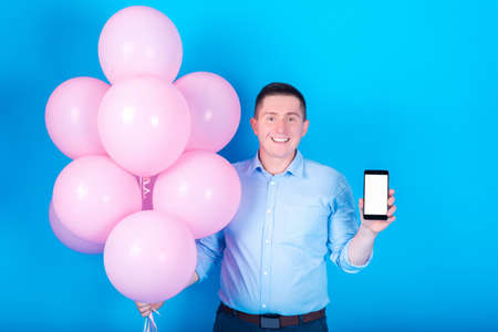 Young handsome smiling man in blue shirt holding pink air balloons and smartphone with white blank...