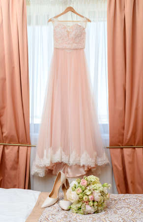 Beautiful lush wedding bouquet of pink and white flowers and bride's shoes on bed with wedding dress on background in bride's bedroom の写真素材