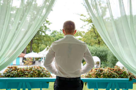 Back view of young man dressed in white shirt and black trousers on porch outdoor, free space. Groom standing on wooden terrace with green nature view. Business man  enjoying the view from balconyの写真素材