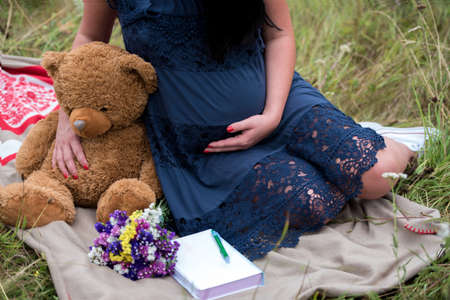 Pregnant woman in dress holding teddy bear to her tummy. Closeup of pregnant woman with teddy bear sitting on blanket on green grass outdoorsの写真素材