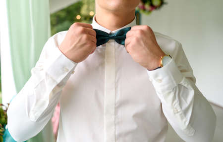 Close-up of businessman or groom holding bow-tie in hands. Man adjusting or correcting emerald bowtie in white shirt. Formal style of clothes. Person ready for wedding or graduationの写真素材