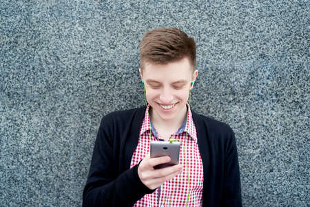 Cheerful happy young man in plaid shirt and jacket listening to music with headphones from mobile...