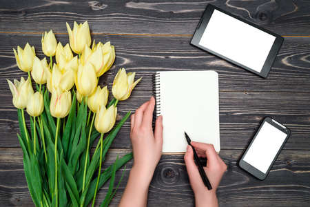 Spring background with yellow tulip flowers, mobile smart pnone with white screen, tablet and woman hands with pen writing in blank notebook, copy space. Top view. Woman's Day (March 8), Mother's Dayの写真素材