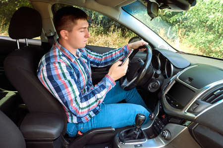 Side view of handsome serious young man in plaid shirt driving his car and using smart phone. Young man with hand on steering wheel using mobile phone while driving. Doing forbidden thingsの写真素材