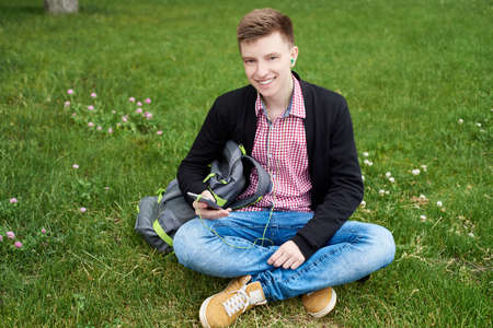 Portrait of young happy smiling man with backpack sitting on green grass lawn and listening to music with headphones from mobile smart phone outdoors, enjoying nature, copy spaceの写真素材