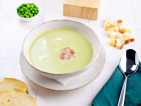 Green pea cream soup with bacon and cream in bowl with croutons on white wooden table. Light summer green pea soup in bowl with bread toastsの写真素材