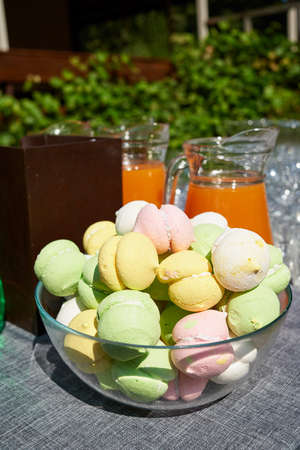 Lot of colorful meringue cookies in glass bowl with orange juice on table outdoors, free space. Delicious homemade meringues. Sweet foodの写真素材