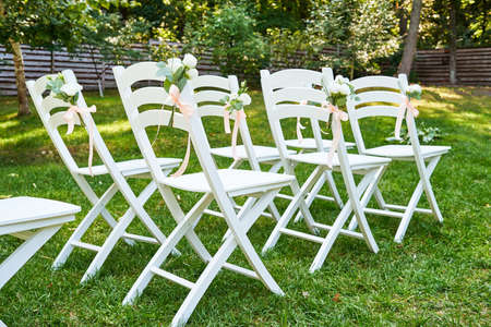 White wedding chairs with fresh flowers on each side of archway outroods, copy space. Empty wooden chairs for guests on green grass in the garden prepared for wedding ceremonyの写真素材