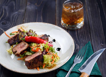 Veal salad with lettuce, carrot, tomato and pepper on white plate on dark wooden background, copy space. Restaurant food, close up. Meat salad and glass of whiskeyの写真素材