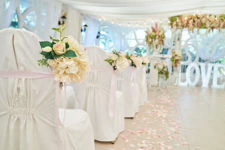 Beautiful flower arrangement on white wedding chairs on each side of archway, selective focus. Empty chairs with white cloth for guests on wedding ceremony, free spaceの写真素材