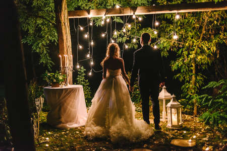 Night wedding ceremony with candles, lanterns and lamps on tree. Bride and groom holding hands on background of bulb lights, back view. Beautiful young couple standing under a tree at nightの写真素材