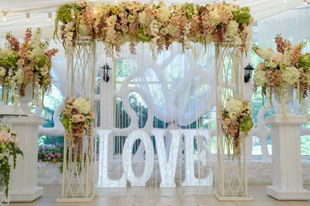 Beautiful white wedding arch for wedding ceremony decorated with flowers, lightbulbs and sign Love with big white letters, copy spaceの写真素材