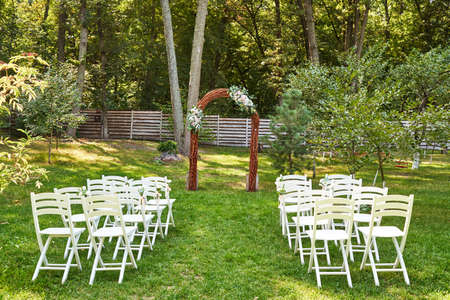 Place for wedding ceremony with wedding arch decorated with flowers and white chairs on each side of archway outdoors, copy space. Empty wooden chairs for guests on green grass in the gardenの写真素材