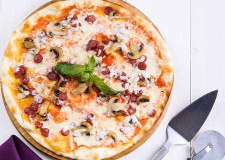 Hot homemade pizza with sausage, mushrooms, mozzarella cheese and basil on wooden board, copy space. Pizza cutter and spatula server. Italian traditional cuisine. Italian food. Top view, flat layの写真素材