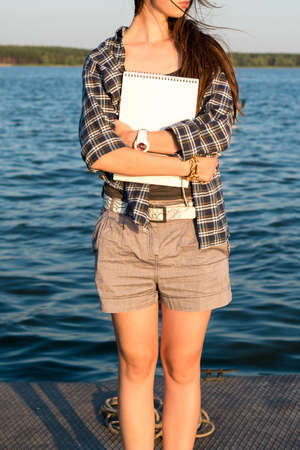 Creative young brunette woman with sketchbook and pencils in hand standing on pier. Vacation, traveling and freedom conceptの写真素材