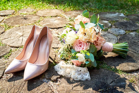 Close up of beige bride shoes with high heels, wedding bouquet and garter on wood background outdoors. Wedding morning preparation. Bride accessories, free spaceの写真素材