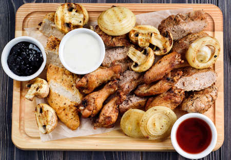 Mixed fresh grilled meat with onion, mushrooms and sauce on wooden cutting board. Assorted delicious barbecue meat with vegetables from summer BBQ. Healthy eating. Top view, flat layの写真素材