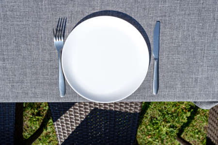 Empty ceramic round plate and cutlery on grey tablecloth outdoors, copy space. Dinner plate setting. White plate with knife and fork on gray backgroundの写真素材