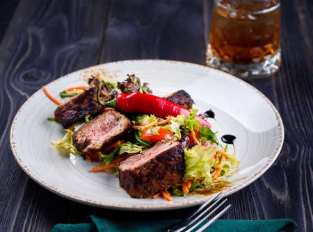 Veal salad with lettuce, carrot, tomato and pepper on white plate on dark wooden background, copy space. Restaurant food, close up. Meat salad and glass of whiskeyの写真素材