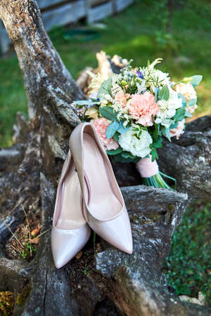 Close up of beautiful wedding bouquet of pink and white roses and beige shiny bride shoes on wooden stump outdoors. Wedding morning preparation. Wedding bride accessories. Bridal bouquet and shoesの写真素材