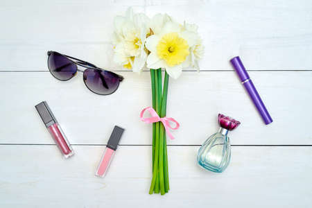Top view of white wooden background with Ñosmetics: mascara, lip gloss and perfume near bouquet of yellow daffodils (narcissus) and sunglasses, copy space. Flat layの写真素材