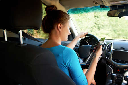 Back view of beautiful young woman driving a car (or standing in traffic congestion) and using mobile smart phone, free space. Business woman sitting in car with cell phoneの写真素材